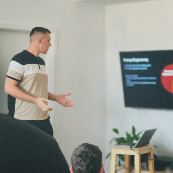 Man in a polo shirt giving a presentation to an audience with a laptop and a screen displaying graphics in the background.