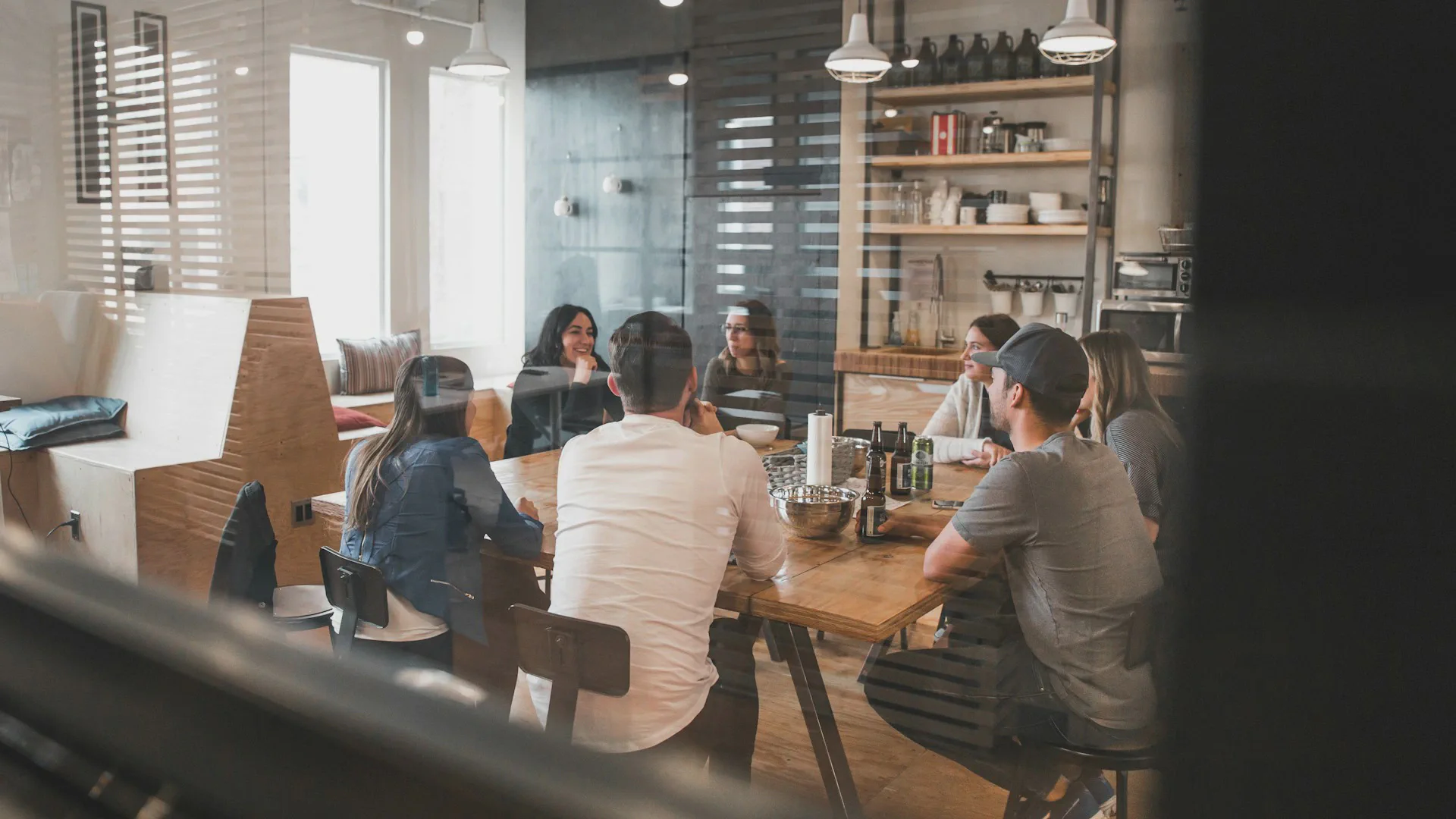 A group of colleagues sitting around a large wooden table in a modern office break room, engaging in conversation.