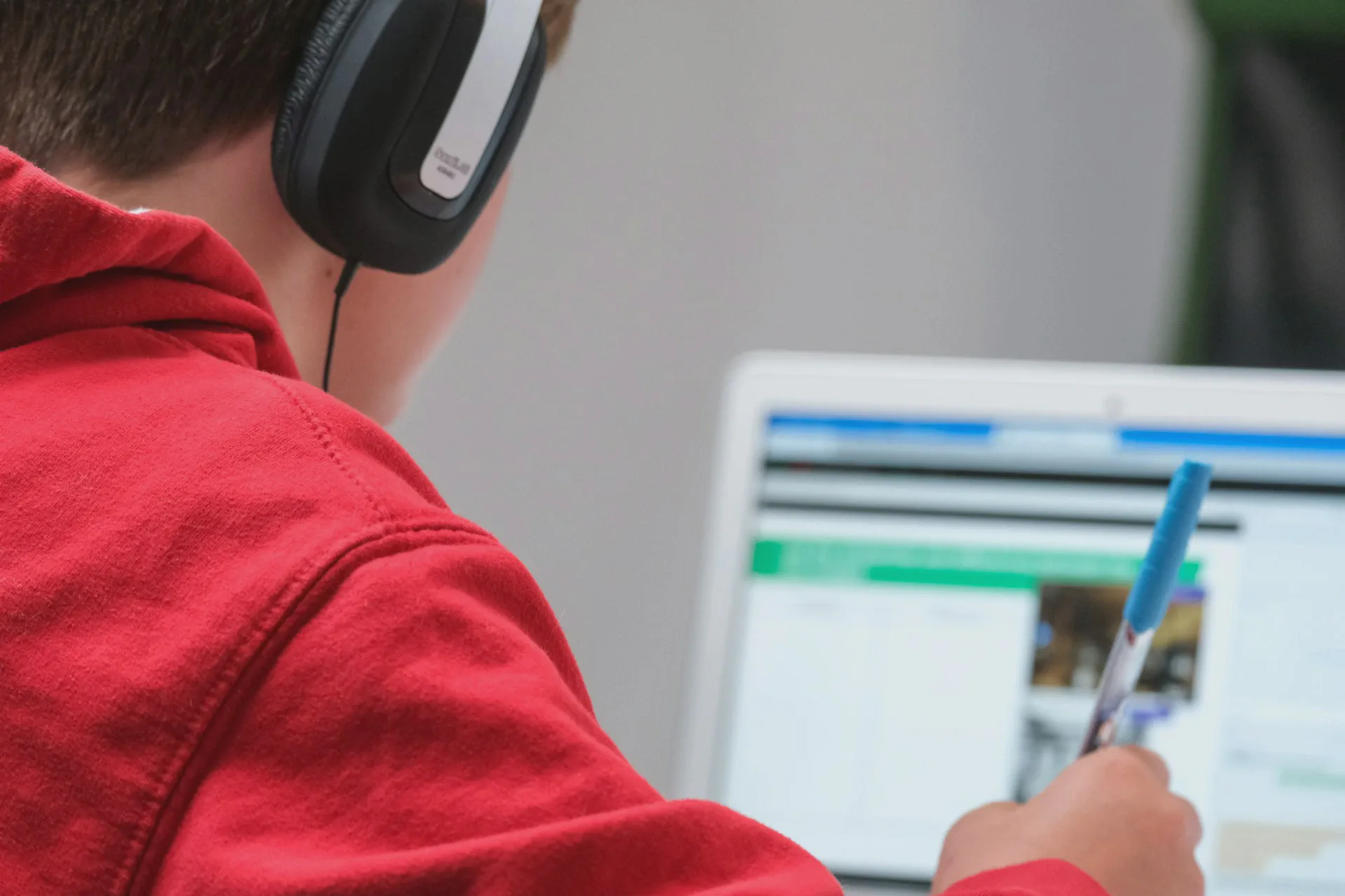 Over-the-shoulder view of a student wearing a red hoodie and headphones, writing notes with a blue pen while looking at an educational platform on a laptop screen.