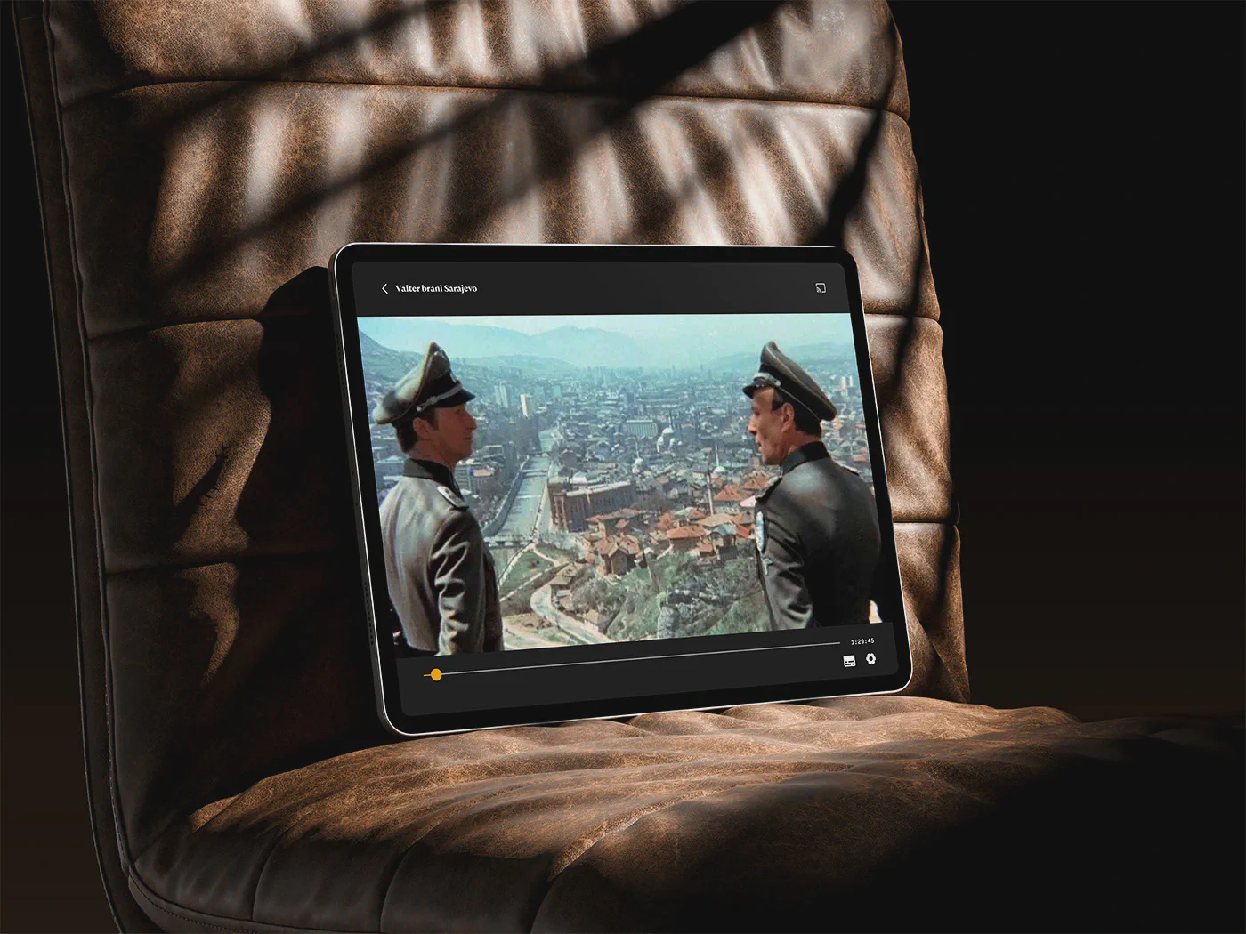 A tablet device resting on a brown leather chair, displaying a streaming video interface playing a scene from the movie 'Valter brani Sarajevo'