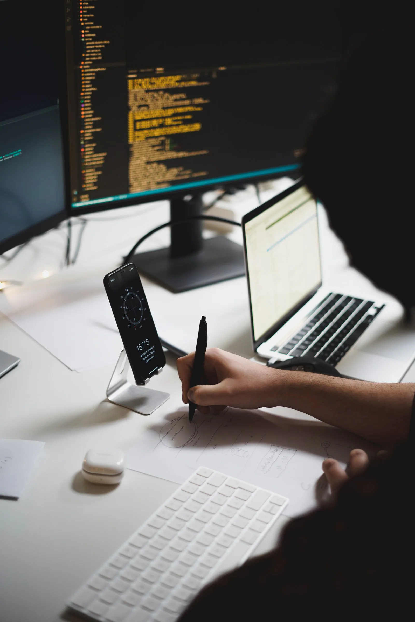 Developer desk with a code editor on screen, a phone on a stand, and a hand sketching a diagram on paper